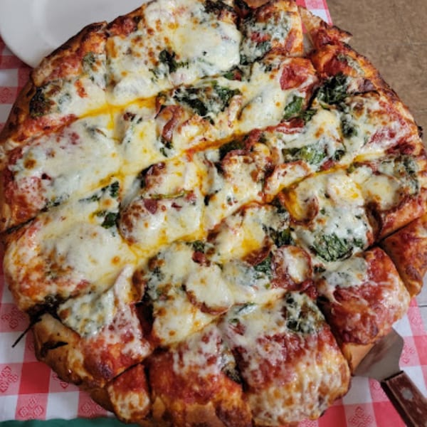 A large, cheesy Pinellas Pizzeria pizza with spinach and pepperoni, sliced on a red and white checkered tablecloth.